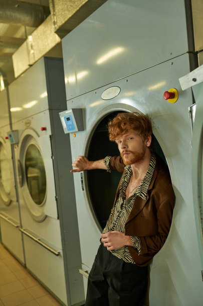 уверенный и стильный молодой человек в куртке позирует в стиральной машине в публичной прачечной - Фото, изображение