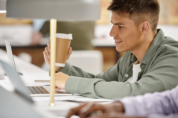 щасливий чоловік тримає каву, щоб піти і використовувати ноутбук, офісне середовище, коворкінг, стартап-проект - Фото, зображення