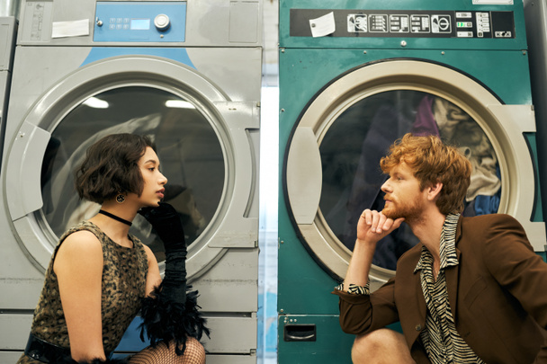side view of trendy multiethnic couple looking at each other near washing machines in public laundry - Photo, Image