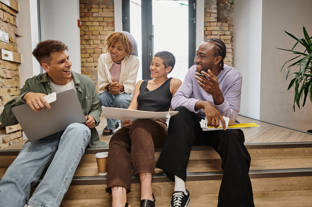 startup, happy diverse team chatting, holding gadgets, sitting on stairs in modern coworking, tech - Φωτογραφία, εικόνα