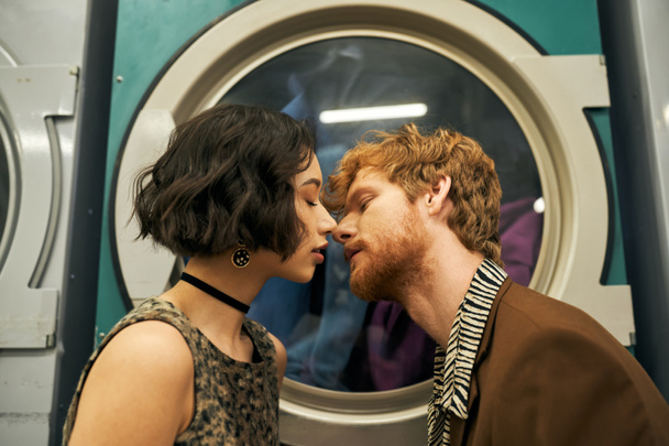 young and stylish multiethnic couple kissing near washing machine in coin laundry - Photo, Image
