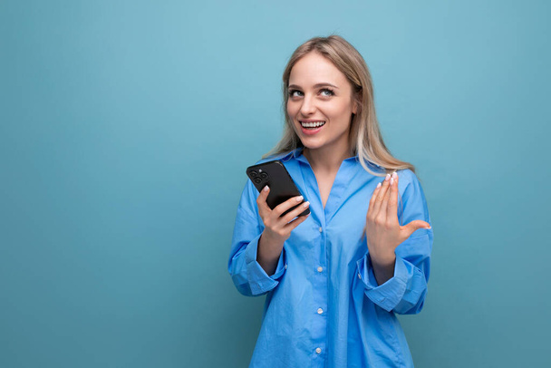 joyful charming blonde girl shocked holding smartphone in hands over isolated blue background. - Photo, Image