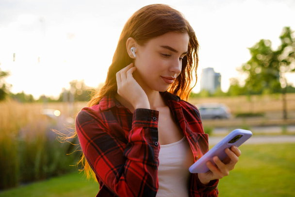 Una joven con auriculares, vistiendo una camisa informal roja y negra, elige cuidadosamente la música en una aplicación en línea en su teléfono inteligente de pie en la calle.  - Foto, Imagen