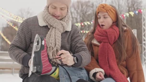 Młody mężczyzna dba o swoją dziewczynę podczas randki na lodowisku, wiąże sznurowadła na łyżwach siedząc na ławce na świeżym powietrzu - Materiał filmowy, wideo