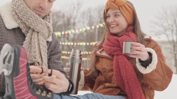Gyönyörű fiatal kaukázusi nő élvezi forró tea termoszból, míg a barátja kötött cipőfűzőt a jégkorcsolya, amelynek romantikus dátum szabadban jégpálya téli ünnepek - Felvétel, videó
