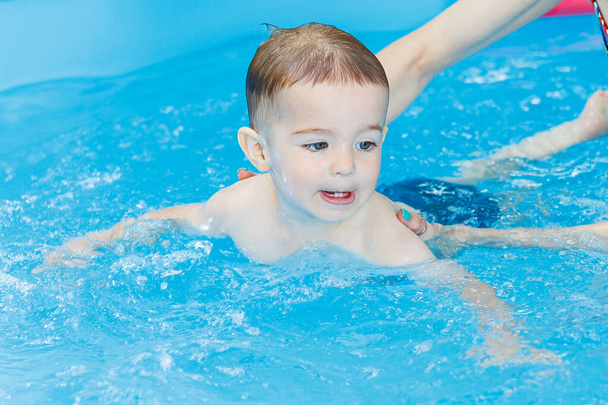 Een 2-jarige jongen leert zwemmen in een zwembad met een coach. Zwemlessen voor kinderen. Zwemschool voor kinderen. Educatieve zwemcursussen voor kinderen - Foto, afbeelding