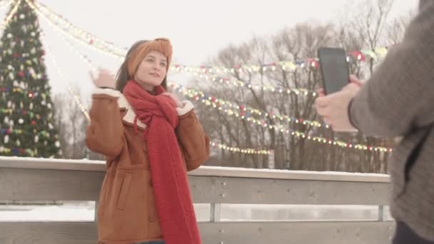 Mladý muž fotí na smartphonu své krásné přítelkyně pózuje pro kameru stojící proti vysokému vánočnímu stromečku s věnci na venkovním kluzišti - Záběry, video
