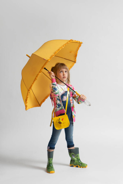 Niña con ropa casual de otoño y botas de goma finge esconderse de la lluvia bajo un paraguas amarillo sobre un fondo blanco - Foto, Imagen