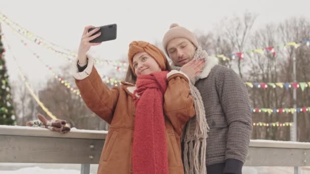 Jovem casal apaixonado tirando selfies no smartphone enquanto está em pé na pista de gelo ao ar livre com árvore de Natal alta lindamente decorada no fundo - Filmagem, Vídeo