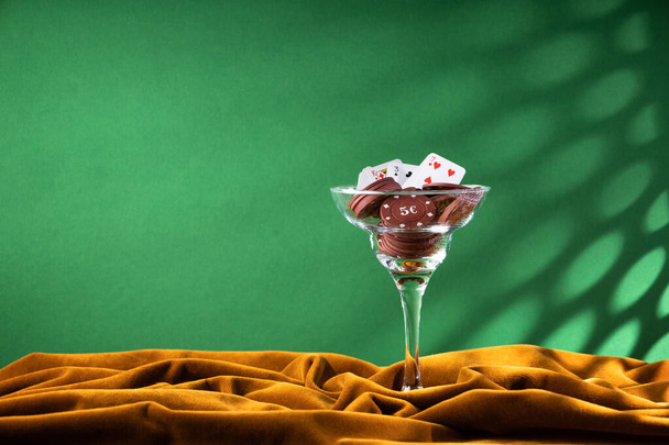 Červené plastové pokerové žetony leží ve skleněné sklenici margarity na hnědém sametovém plátně. Fanoušek malých hracích karet na okraji sklenice, jako koktejl dekorace. Boční pohled - Fotografie, Obrázek