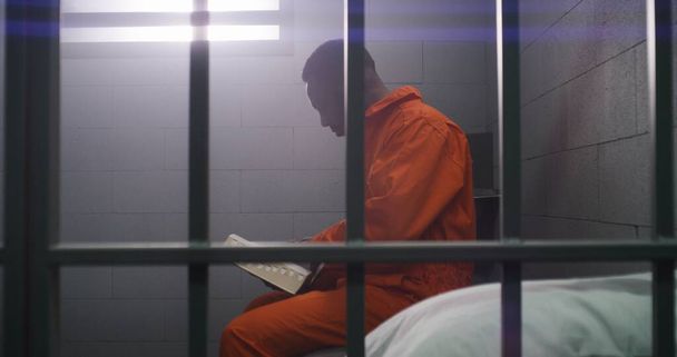 Prisionero afroamericano en uniforme naranja se sienta en la cama tras las rejas, lee la Biblia en la celda de la prisión. Hombre criminal cumple pena de prisión por delito en la cárcel. Centro de detención o correccional. - Foto, imagen