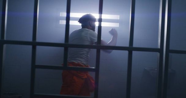 Un prisionero afroamericano con uniforme naranja hace boxeo en la celda. El carcelero camina por el pasillo de la prisión. Recluso cumple pena de prisión por delitos en centro de detención o correccional. - Foto, Imagen