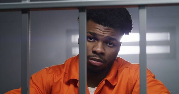 Afro-Amerikaanse man in oranje uniform kijkt naar camera en kneedt de nek in de cel. Gevangene zit gevangenisstraf uit. Crimineel in een gevangenis of detentiecentrum. Portretweergave. - Foto, afbeelding