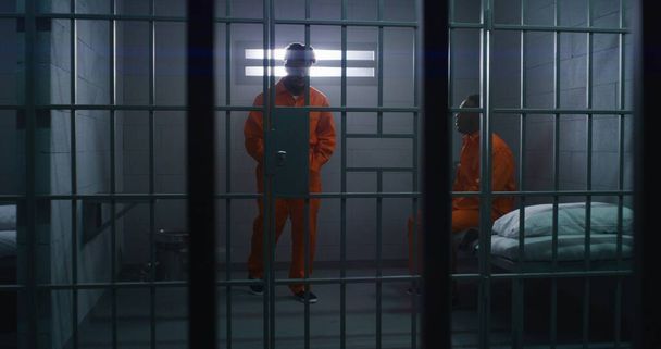 Twee Afro-Amerikaanse gevangenen in oranje uniformen praten. Een man zit op bed, een andere loopt in de cel. Mannen zitten gevangenisstraf uit voor misdaden in de gevangenis, het detentiecentrum of de gevangenis. - Foto, afbeelding