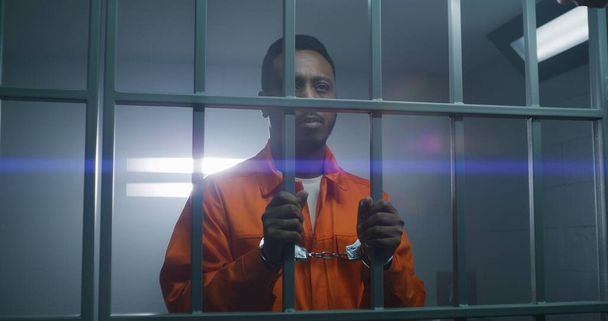 Афроамериканский заключенный в оранжевой форме держит руки в наручниках на тюремных решётках и смотрит в камеру. Виновный преступник отбывает тюремный срок. Печальный убийца в исправительном учреждении. - Фото, изображение