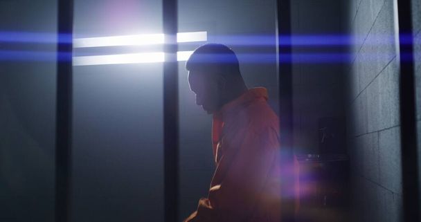 Depressed African American man in orange uniform stands from jail bed, walks at prison cell and looks at barred window. Sad prisoner serves imprisonment term. Murderer in gloomy correctional facility. - Photo, Image