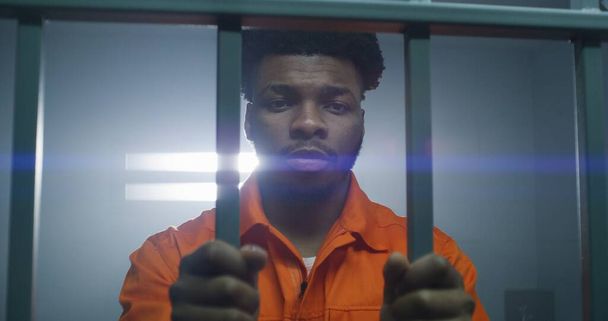 Afro-Amerikaanse man in oranje uniform houdt handen in handboeien op cel bars. Depressieve moordenaar zit gevangenisstraf uit. Schuldige gevangene in een gevangenis of detentiecentrum. - Foto, afbeelding