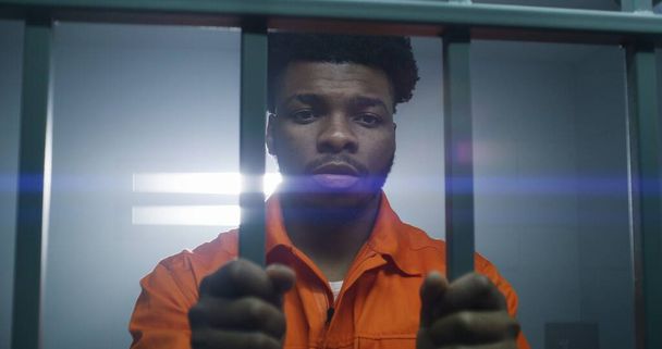 African American man in orange uniform keeps hands in handcuffs on jail cell bars. Depressed murderer serves imprisonment term in prison. Guilty prisoner in correctional facility or detention center. - Photo, Image