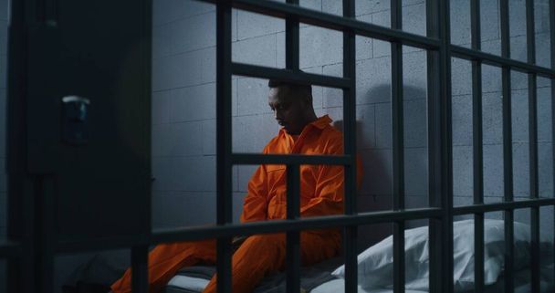 Guilty African American man in orange uniform sits on prison bed and thinks about freedom. Gloomy criminal in correctional facility or detention center. Prisoner serves imprisonment term in jail cell. - Photo, Image