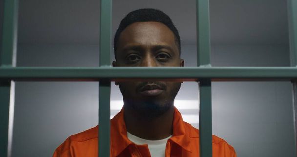 African American man in orange uniform keeps hands in handcuffs on jail cell bars and looks at camera. Sad criminal serves imprisonment term in prison. Prisoner in correctional facility. - Photo, Image