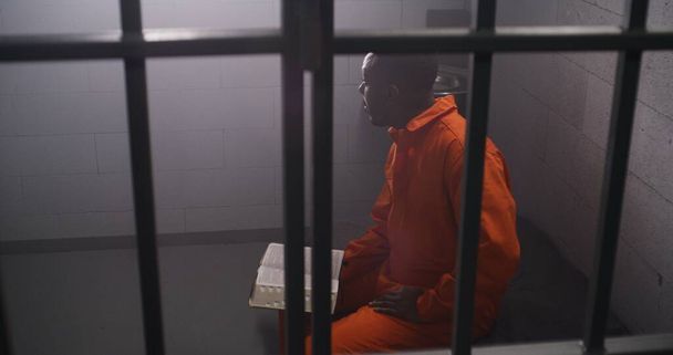 African American prisoner in orange uniform sits on bed behind bars, reads Bible in prison cell. Male criminal serves imprisonment term for crime in jail. Detention center or correctional facility. - Photo, Image