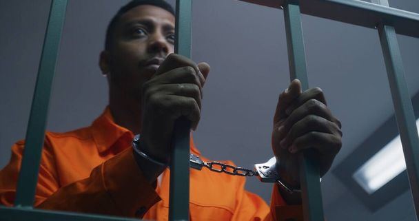 Afroamerykanin w pomarańczowym mundurze trzyma ręce w kajdankach na więziennych kratach. Przygnębiony morderca odsiaduje wyrok więzienia. Więzień winny w zakładzie karnym lub areszcie. - Zdjęcie, obraz