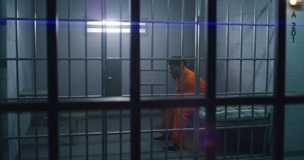 Een man in oranje uniform geeft eten van serveerwagen tot gevangene in de cel. Depressieve crimineel zit op bed, eet eten, zit gevangenisstraf uit voor misdaad. Gevangenis of gevangenis. - Foto, afbeelding