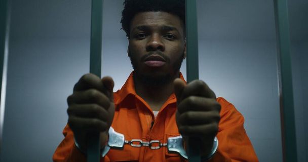 Un Afro-Américain en uniforme orange garde les mains menottées sur les barreaux de prison. Un meurtrier déprimé purge une peine d'emprisonnement. Prisonnier coupable dans un établissement correctionnel ou un centre de détention. - Photo, image