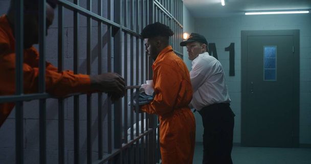 Warden brings new prisoner in jail cell and takes off his handcuffs. African American criminals serve imprisonment term in correctional facility or detention center. Guilty murderers in prison cells. - Photo, Image