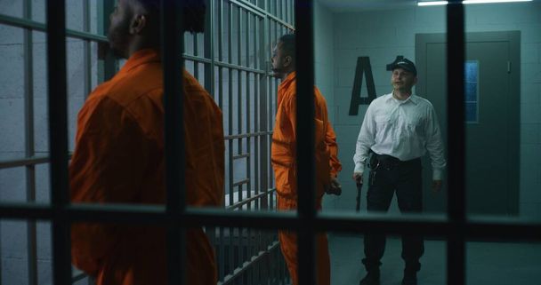 Gefängnismitarbeiterin mit Polizeiknüppel geht auf Gefängnisflur und schikaniert Gefangene. Afroamerikanische Kriminelle stehen mit den Händen hinter der Zelle. Mörder verbüßen Haftstrafe in Haftanstalt. - Foto, Bild