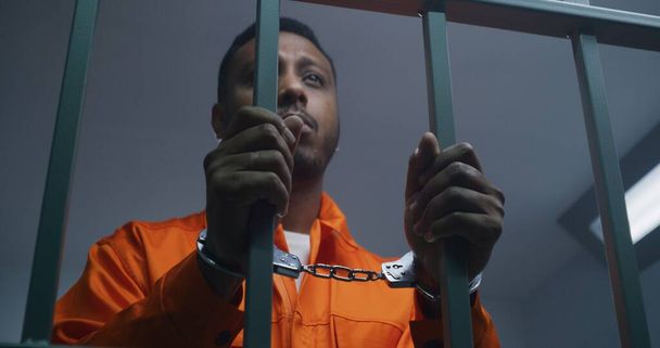African American man in orange uniform keeps hands in handcuffs on jail cell bars. Depressed murderer serves imprisonment term in prison. Guilty prisoner in correctional facility or detention center. - Photo, Image