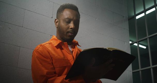 African American prisoner in orange uniform sits in prison cell, reads Bible. Male criminal serves imprisonment term for crime in jail. Offender in detention center or correctional facility. Portrait. - Photo, Image