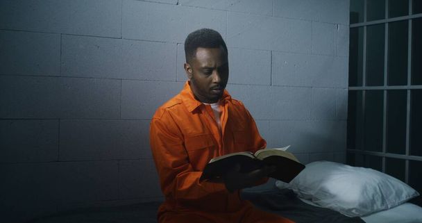 Ο Αφρο-Αμερικανός κρατούμενος με πορτοκαλί στολή κάθεται στο κελί της φυλακής και διαβάζει τη Βίβλο. Άντρας εγκληματίας εκτίει ποινή φυλάκισης για έγκλημα στη φυλακή. Παραβάτης σε κρατητήριο ή σωφρονιστικό ίδρυμα. Πορτρέτο. - Φωτογραφία, εικόνα
