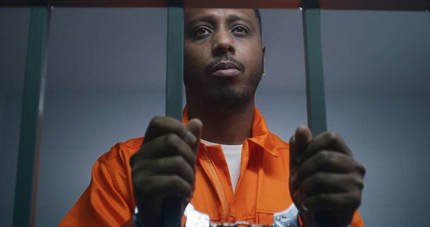 Afroamerikkalainen mies oranssissa univormussa pitää käsiraudoissa sellibaareja. Masentunut murhaaja istuu vankeusrangaistusta. Syyllinen vanki vankilassa tai säilöönottokeskuksessa. - Valokuva, kuva