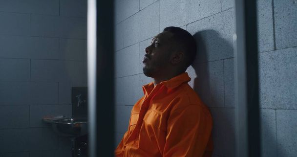 Un afroamericano culpable con uniforme naranja se sienta en la cama de la prisión y piensa en la libertad. Delincuente sombrío en correccional o centro de detención. Prisionero cumple pena de prisión en celda. - Foto, imagen