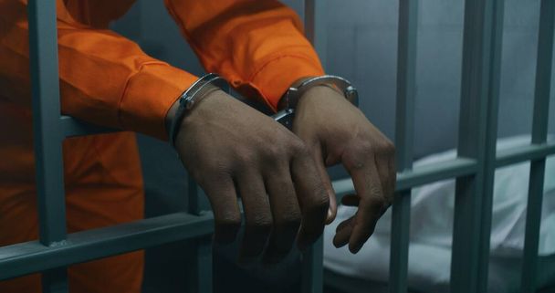 Close up shot of hands in handcuffs leaning on prison cell bars. Criminal serves imprisonment term in correctional facility or detention center. African American prisoner in orange uniform in jail. - Photo, Image