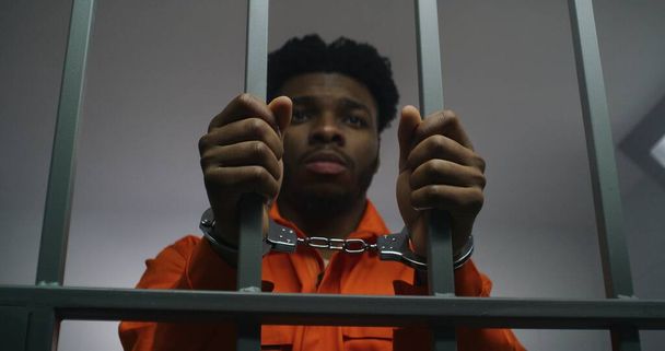 Afroamerikaner in orangefarbener Uniform halten die Hände in Handschellen an Gittern von Gefängniszellen. Depressiver Mörder verbüßt Gefängnisstrafe Schuldiger Häftling in Justizvollzugsanstalt oder Haftanstalt. - Foto, Bild