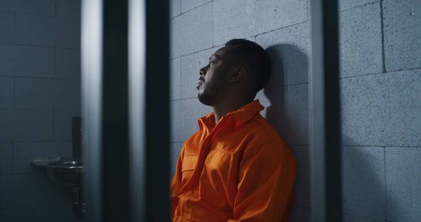 Un afroamericano culpable con uniforme naranja se sienta en la cama de la prisión y piensa en la libertad. Delincuente sombrío en correccional o centro de detención. Prisionero cumple pena de prisión en celda. - Foto, Imagen