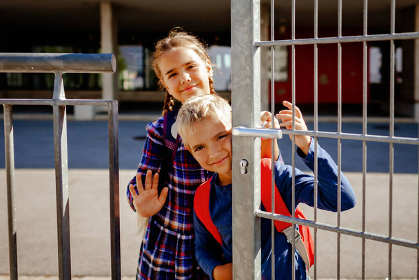 Portrait of happy siblings with backpacks, primary school kids, looking out from the gate near the school at first day, study and education, reunion after vacations, lifestyle. - Photo, Image