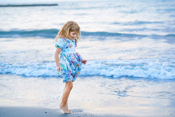 Niño feliz, niña preescolar en vestido corriendo y saltando en las olas durante las vacaciones de verano en la playa tropical exótica al atardecer. Viaje familiar en la costa del océano - Foto, Imagen
