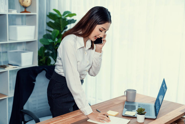 Young enthusiastic businesswoman talking on business call and writing notes on her laptop as multitask office lady. Female employee writing business tasks while talking on phone call with clients. - Photo, Image