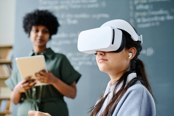 Student in VR glasses sitting at lesson with teacher explaining material in background - Photo, Image
