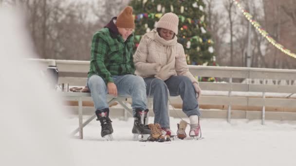 Fröhliches multiethnisches lesbisches Paar, das sich umarmt, während es auf einer Bank an der Eisbahn sitzt und seine Schlittschuhe anzieht - Filmmaterial, Video