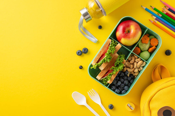 Ernährungsbewusstes Schulessenkonzept. Ansicht von oben mit umweltbewusster Lunchbox mit Bio-Leckereien, Trinkflasche, Besteck, Kunstgegenständen, verspieltem Rucksack auf gelber Oberfläche mit Platz für Text oder Werbung - Foto, Bild
