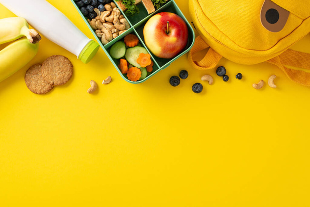 Odżywianie spełnia kreatywność w tej koncepcji posiłek szkolny: Widok z góry pudełko lunchbox zapakowane zdrowych smakołyków, butelka jogurtu, i zabawny plecak na żółtej powierzchni - Zdjęcie, obraz