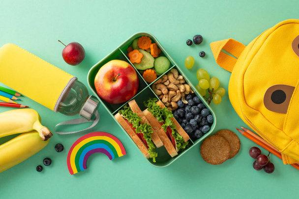 Prioritize health in classroom - top view of lunchbox filled with fresh food, cookies, water, stationery, color pencils, rainbow plasticine, funny backpack on teal background, perfect for promotion - Photo, Image