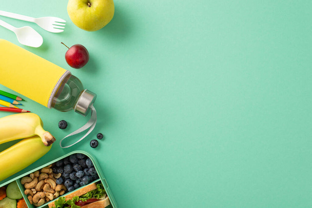 Illuminez la journée scolaire avec un déjeuner sain : Vue du dessus de la lunchbox en plastique contenant des aliments naturels, pomme, prune, myrtilles, noix de cajou, sandwich, bouteille d'eau, couverts, crayons de couleur sur fond turquoise - Photo, image