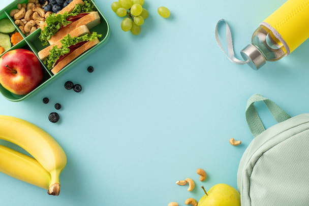 Eine ernährende Schulpausenszene von oben, die eine Lunchbox mit belegten Broten, Obst, Beeren, Wasserflasche und Rucksack vor blauem Hintergrund zeigt, perfekt für Text oder Werbung. - Foto, Bild