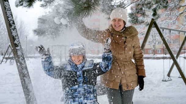 Mère rieuse avec son fils secouant une branche d'arbre et appréciant tomber de la neige sur la tête. Fun et joie pendant les vacances d'hiver, enfants jouant à l'extérieur, activité dans la neige - Photo, image
