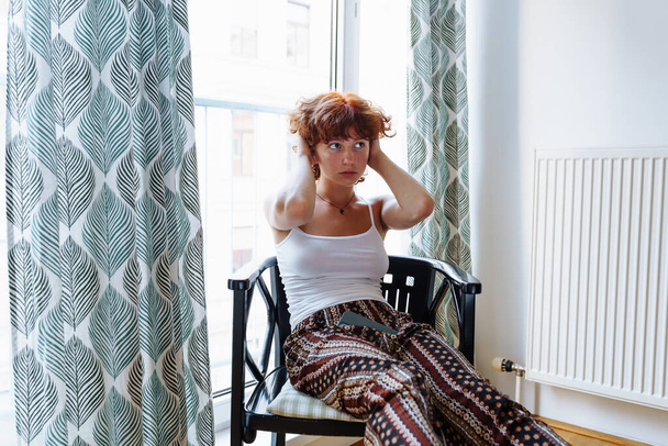 Retrato atraente adolescente ruiva, em roupas de casa, sentado na cadeira na sala de estar perto de uma grande janela. - Foto, Imagem